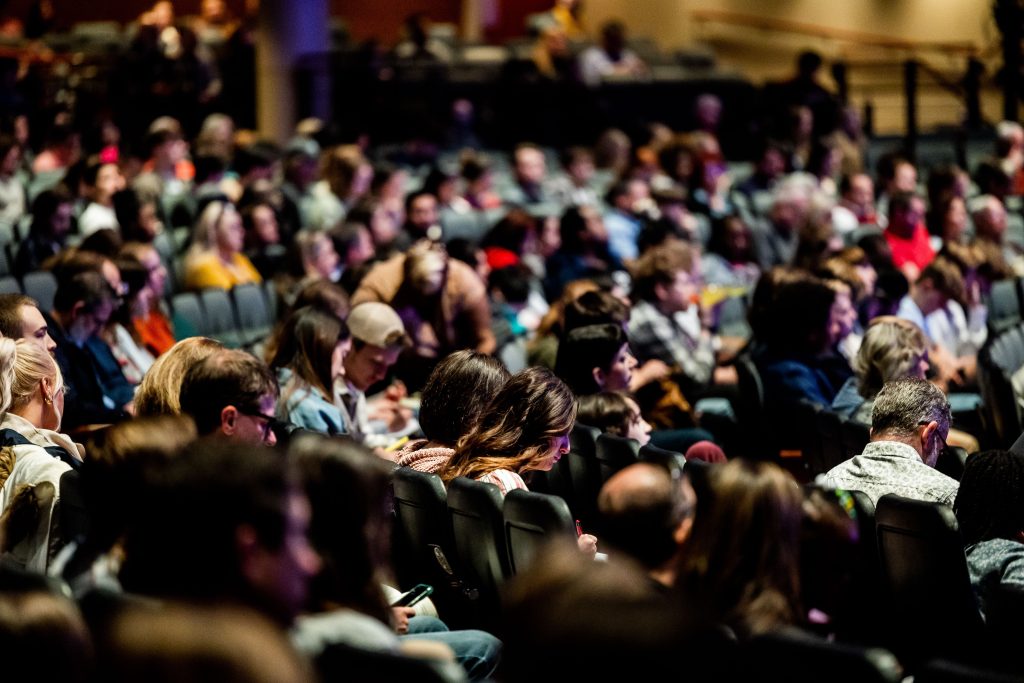 A diverse crowd seated in an auditorium, engaged in an indoor event.